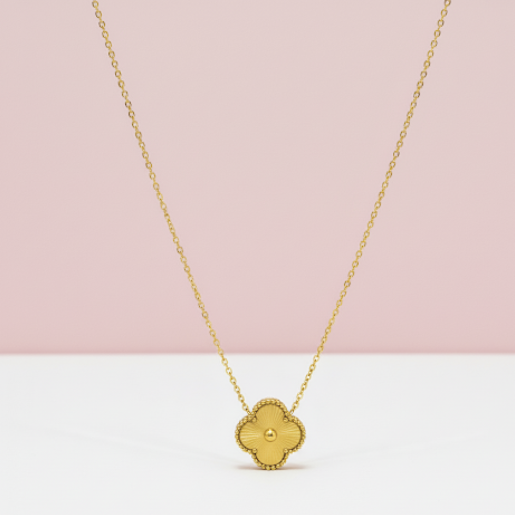 Gold necklace with a clover-shaped pendant on a pink and white background